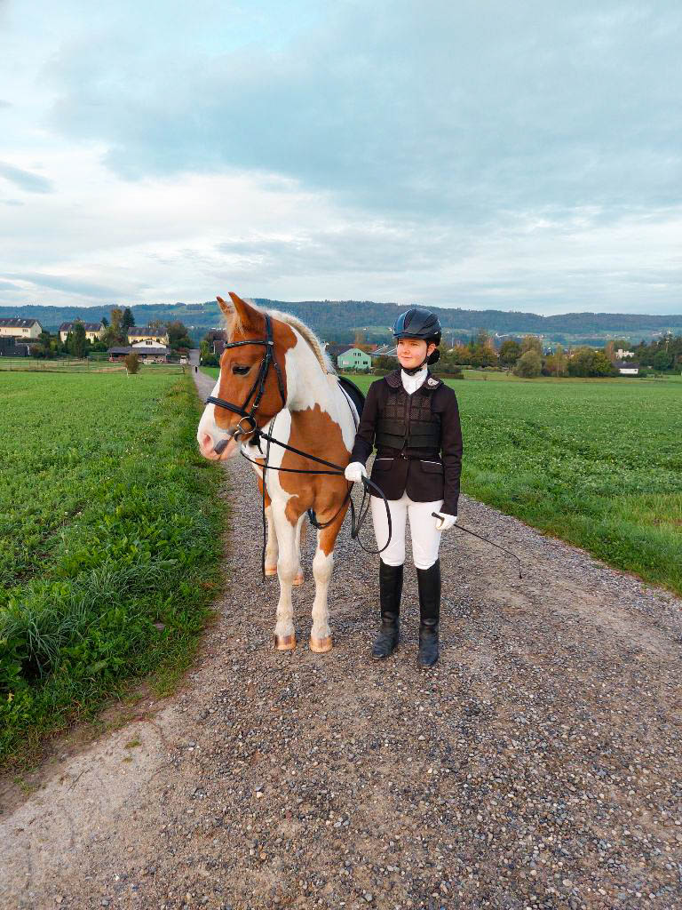 Reiterin in Turnierkleidung steht mit geschecktem Pferd auf Kiesweg im Grünen.